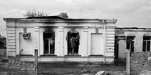 Damaged white building with broken windows and bullet holes, set against a cloudy sky.