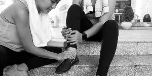 Two women on outdoor steps; one adjusts the other's ankle while both wear athletic clothing and towels.