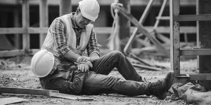 A construction worker assists an injured colleague on the ground, both wearing hard hats and work attire.