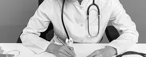 A doctor in a white coat with a stethoscope writes notes at a desk.