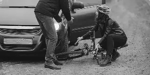 A woman in a helmet kneels beside a bicycle, while a man assists her near a parked car on a gravel road.