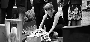 Woman in black kneels at a grave, placing flowers, surrounded by mourners in a cemetery.